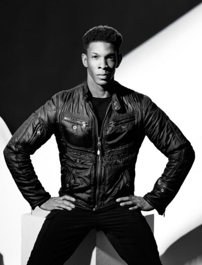 Another monochrome portrait of the same dancer in the leather jacket, now posed with his hands on his hips. The lighting again creates striking geometric shadows across the background, reinforcing the graphic composition. His confident stance gives the image a strong fashion-editorial energy.