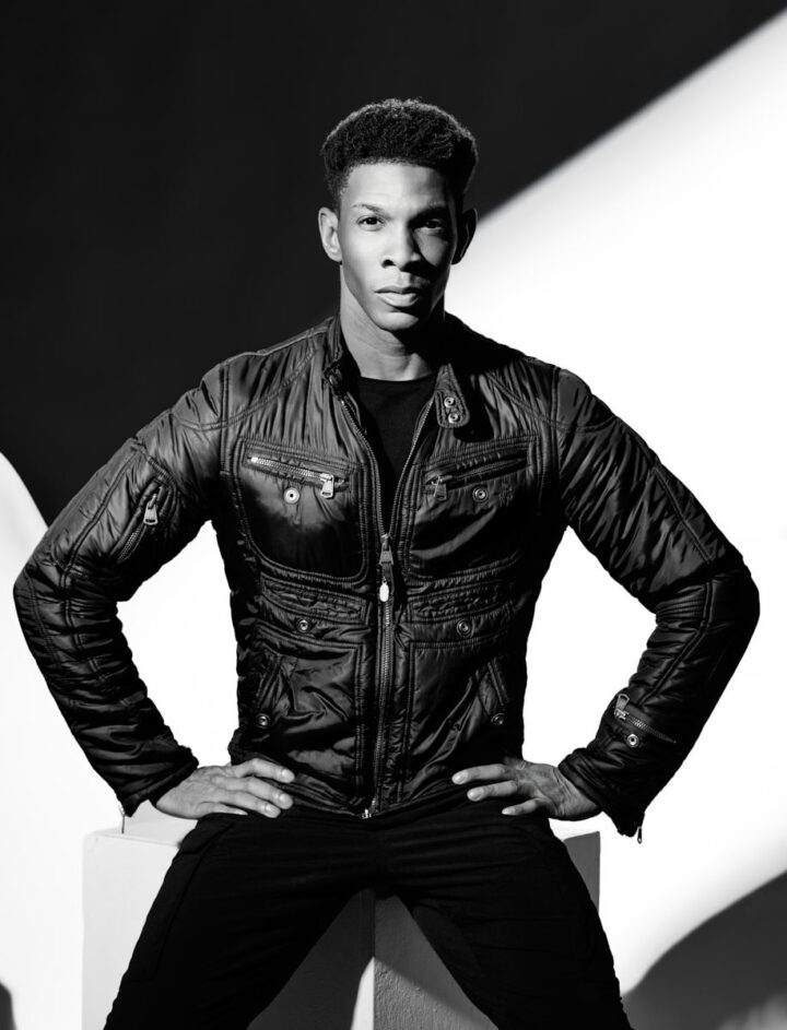 Another monochrome portrait of the same dancer in the leather jacket, now posed with his hands on his hips. The lighting again creates striking geometric shadows across the background, reinforcing the graphic composition. His confident stance gives the image a strong fashion-editorial energy.