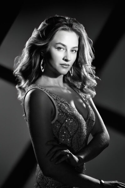 A glamorous black-and-white portrait of the same woman styled in a shimmering evening dress. Her hair is styled in soft waves, and she wears statement earrings that catch the light. She poses with her arms crossed gracefully while dramatic lighting creates bold shadows in the background, evoking a classic Hollywood-inspired aesthetic.