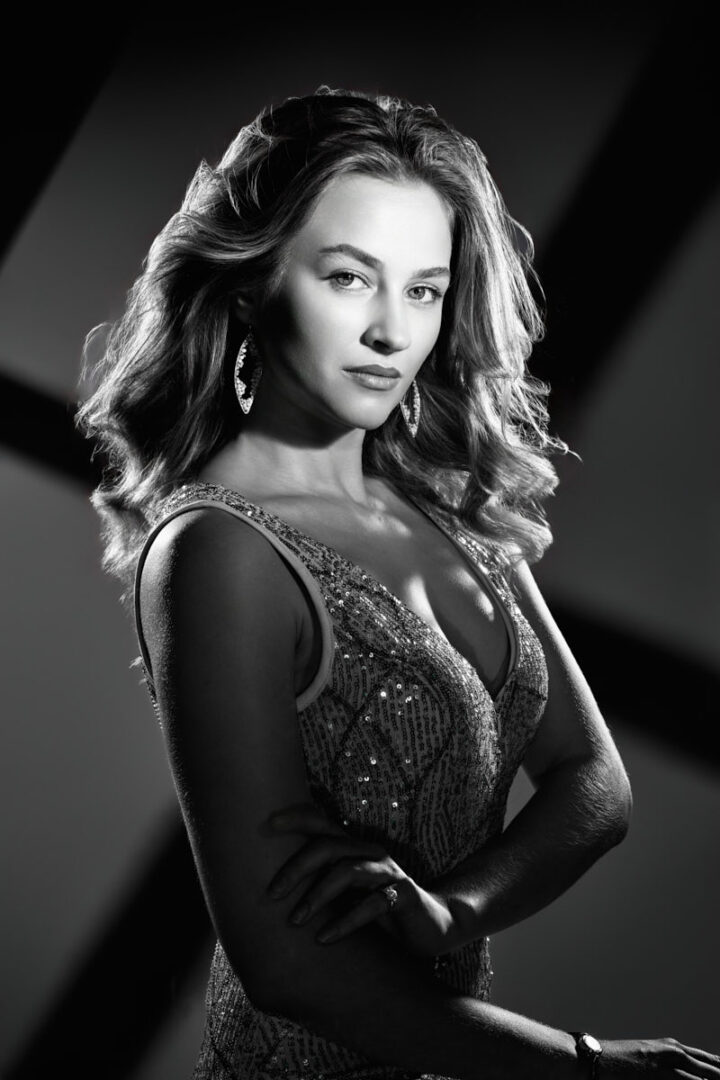 A glamorous black-and-white portrait of the same woman styled in a shimmering evening dress. Her hair is styled in soft waves, and she wears statement earrings that catch the light. She poses with her arms crossed gracefully while dramatic lighting creates bold shadows in the background, evoking a classic Hollywood-inspired aesthetic.