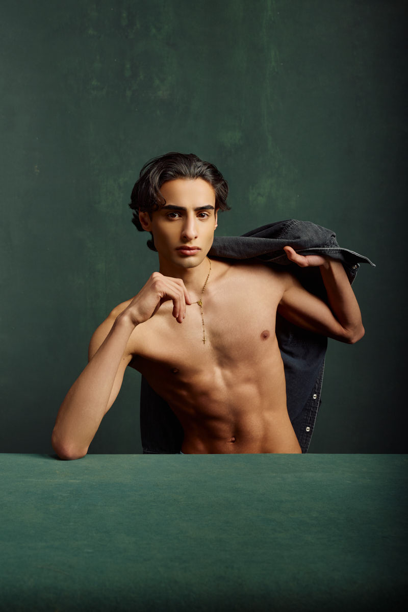 Giorgio leaning forward with a denim jacket over his shoulder, showcasing a strong editorial pose against a textured green backdrop.