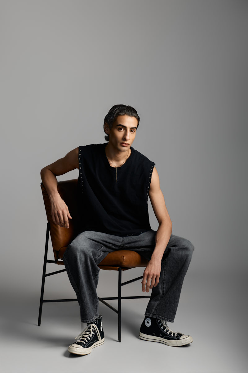 Giorgio seated on a leather chair wearing a black sleeveless shirt, gray jeans, and Converse sneakers in a contemporary fashion portrait.