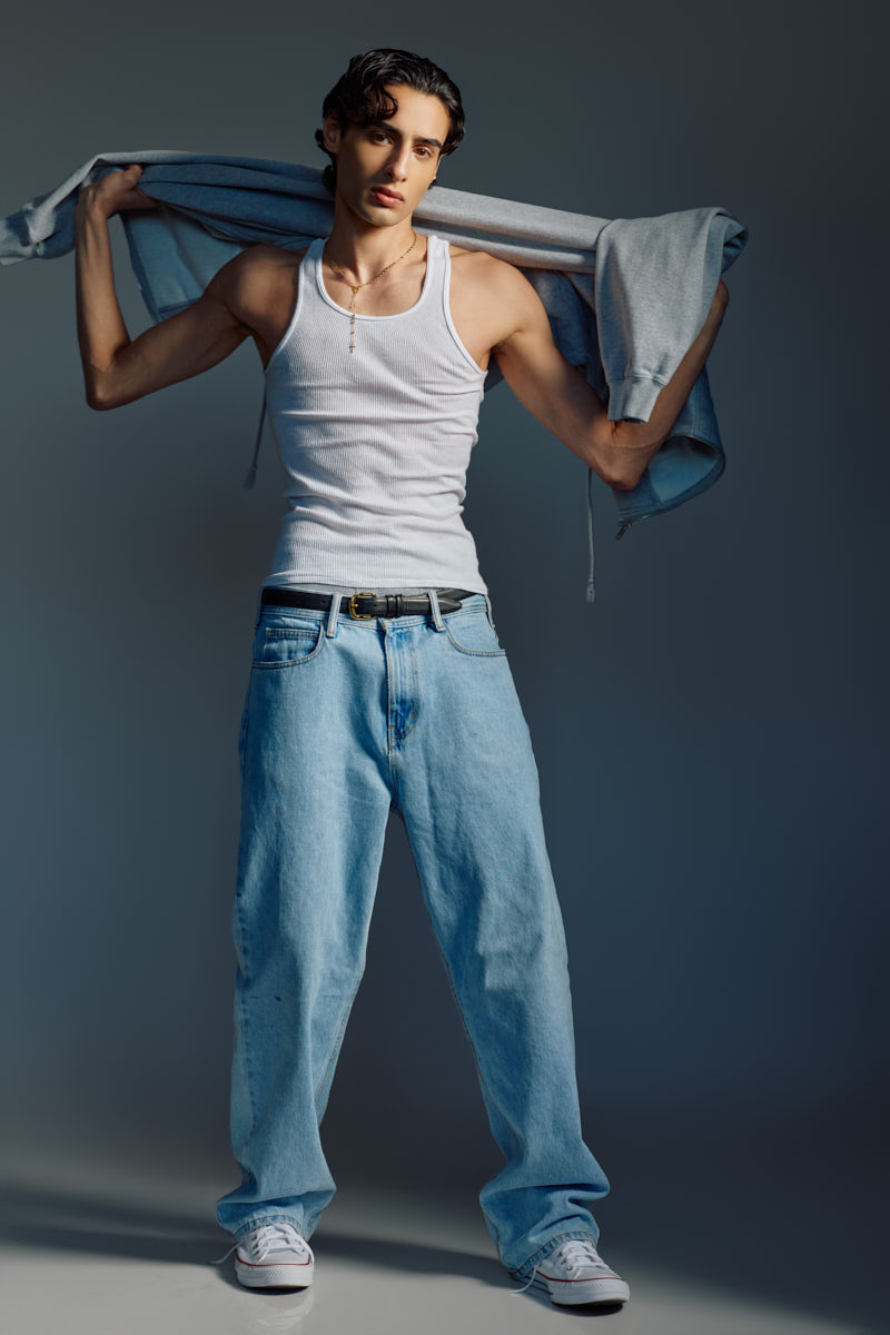 Full-length portrait of Giorgio wearing a white tank top and relaxed-fit jeans while holding a hoodie over his shoulders.