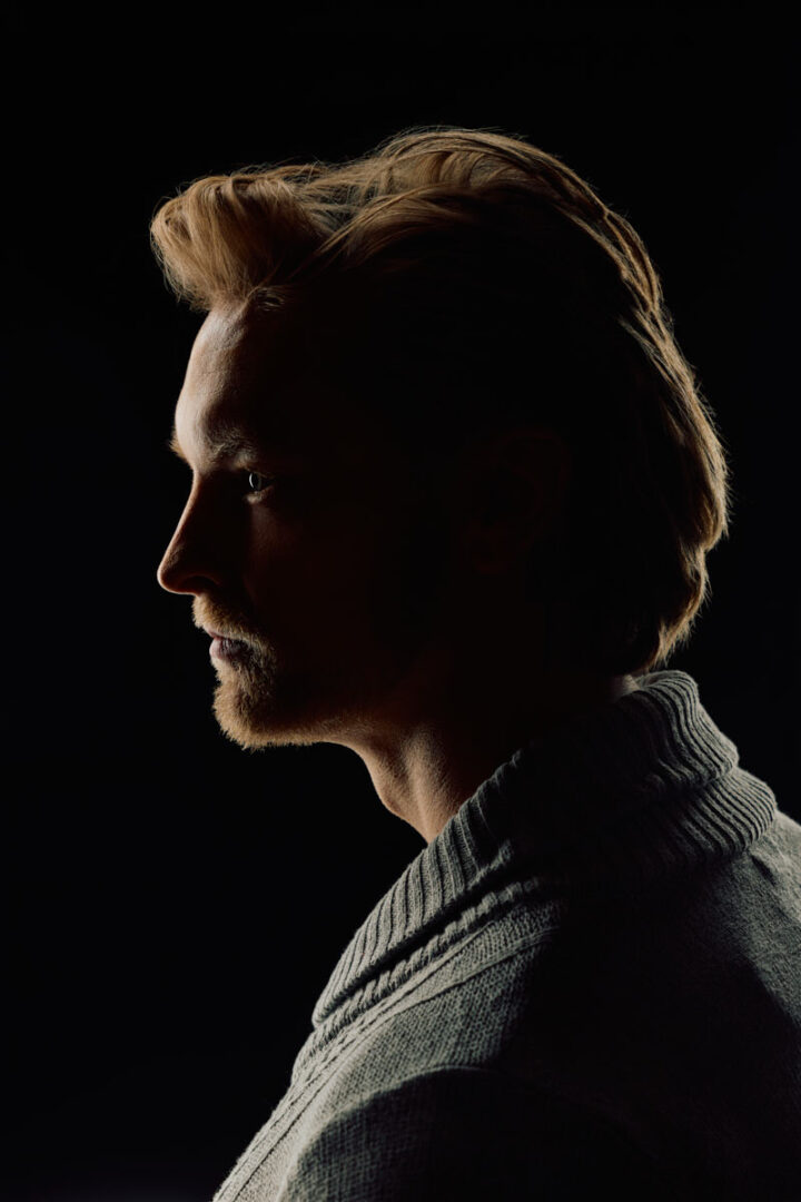 The final portrait shows Nick in dramatic profile against a black background. A narrow rim of light outlines his forehead, nose, beard, hair, and shoulder. Most of his face remains in shadow, creating a striking low-key portrait that emphasizes shape and texture.