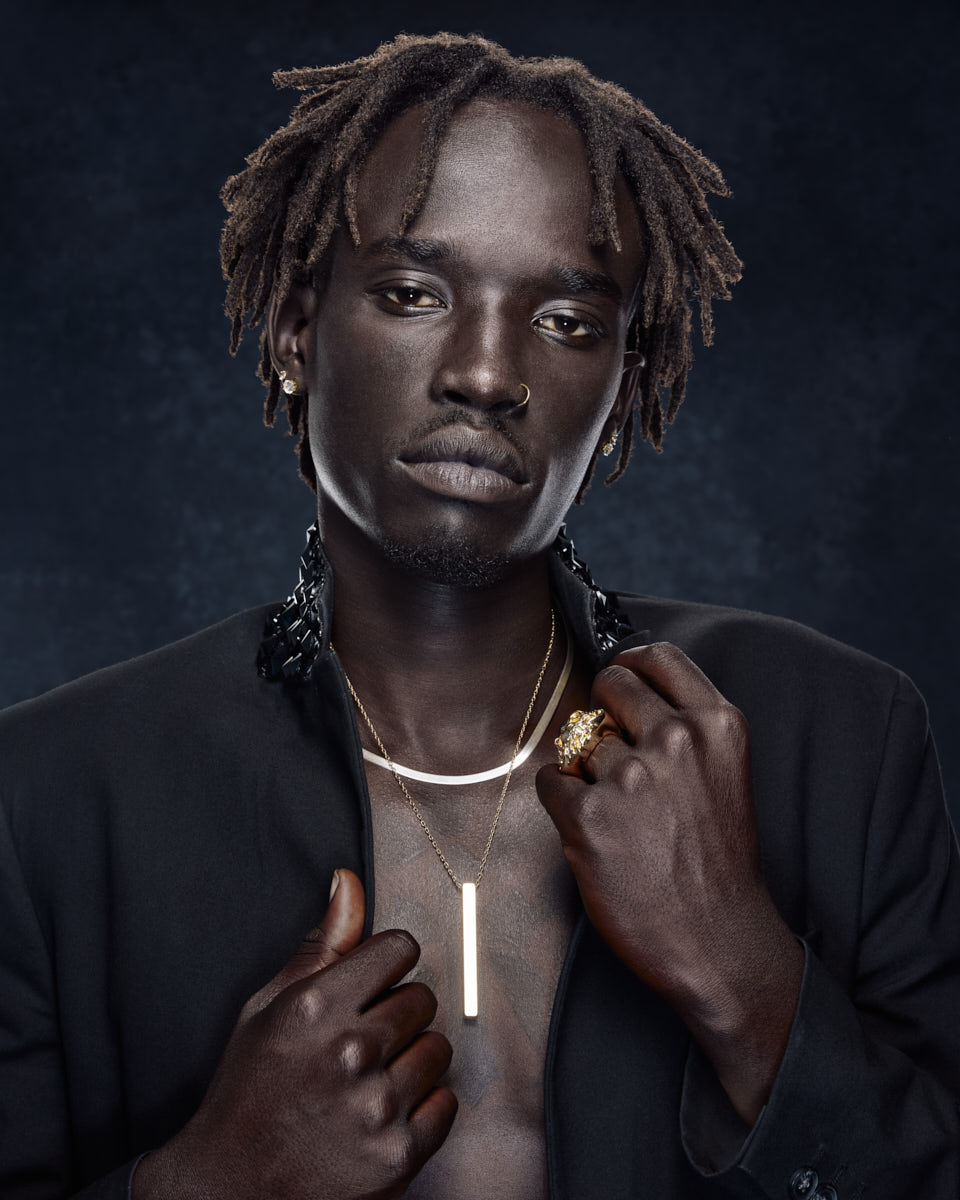 A close-up portrait of Jada wearing a black outfit with gold jewelry. The background is dark, highlighting his face and upper body.