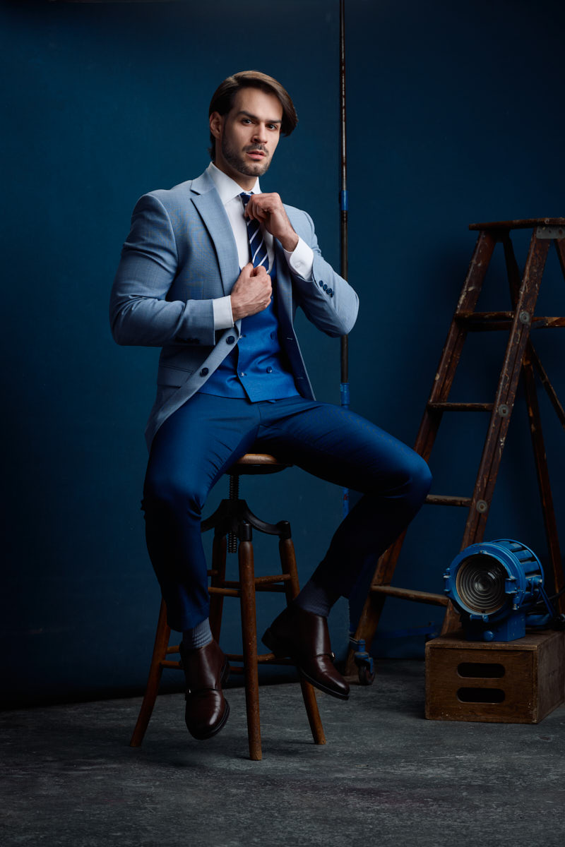 Athletic model photographer Chicago portrait of Jamie adjusting his tie in a blue three-piece suit beside vintage studio props.