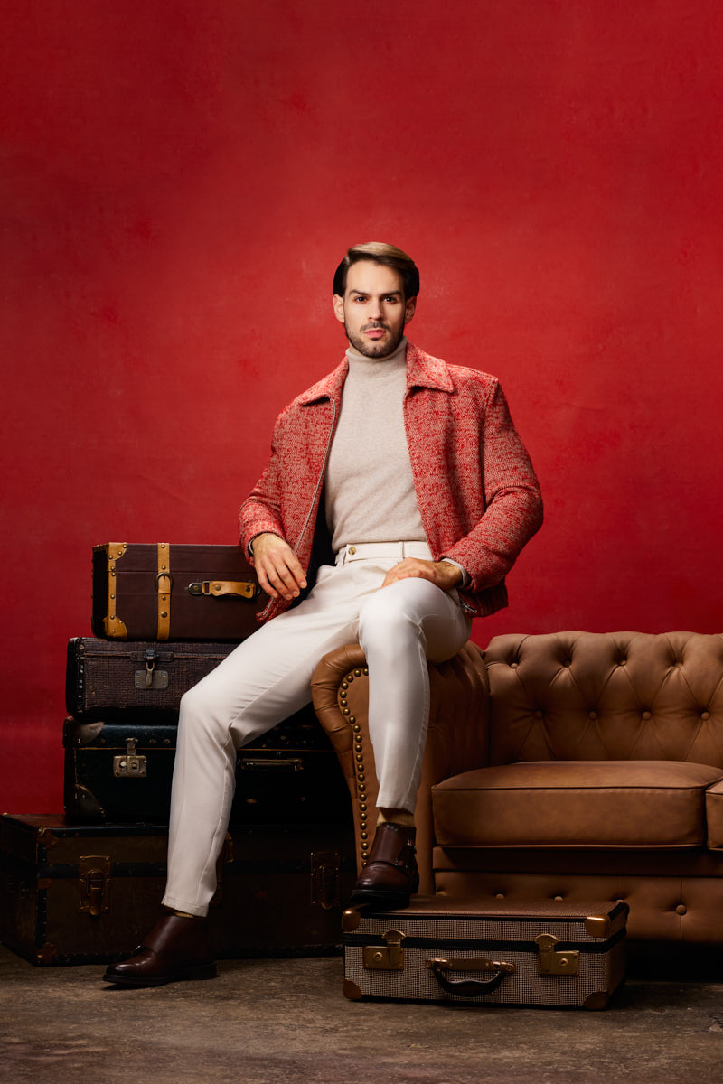 Jamie poses between stacked vintage suitcases and a tufted leather sofa against a textured red background.