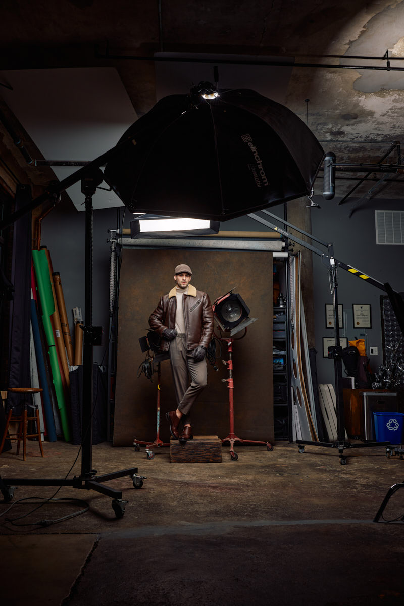 Behind-the-scenes studio portrait showing Jamie in heritage menswear beneath a large overhead flash modifier.