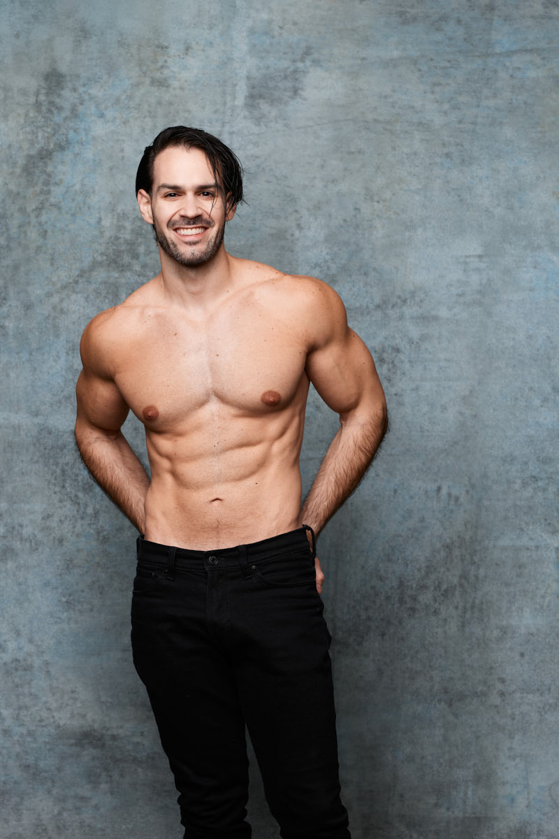 Jamie smiles shirtless against a blue-gray background in an approachable commercial athletic modeling portrait.