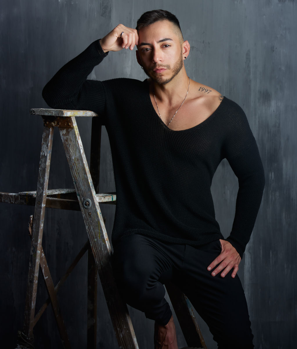 Gael rests an arm on a ladder while wearing a black V-neck top against a gray backdrop.