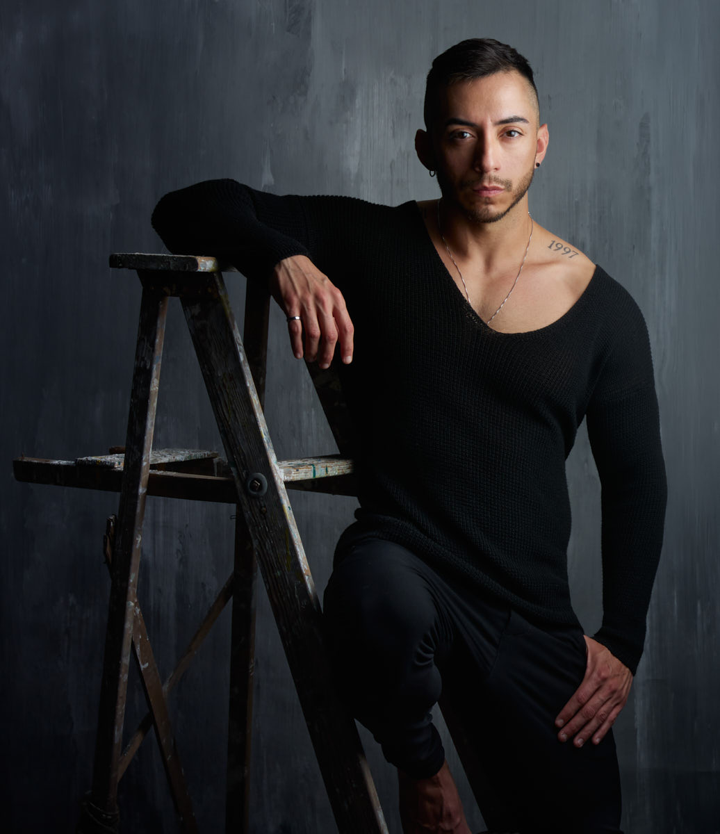 Gael leans on a ladder in a black shirt and trousers in a studio portrait.